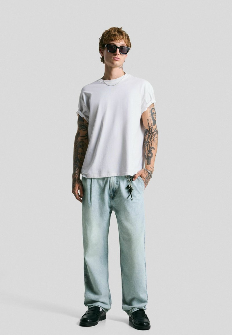 Witte oversized t-shirt met opgerolde mouwen, lichtblauwe baggy jeans, zwarte zonnebril en zwarte instappers met een sleutelhanger-detail.