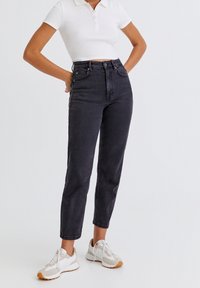 Jeans de mezclilla negros de alta cintura con un corte ajustado, cuentan con un diseño de cinco bolsillos y cierre frontal de botón, combinados con una polo blanca de manga corta.