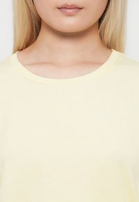 Nærbilde av en persons nedre ansikt og nakke iført en lys gul t-skjorte med rund hals, rett og blondt hår.