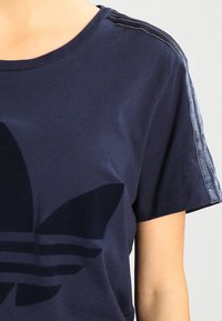 Marineblauwe T-shirt met korte mouwen, voorzien van een groot zwart logo-ontwerp en subtiele contrasterende strepen langs de schouders.
