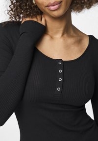 Black long-sleeve top in ribbed fabric features a rounded neckline and five silver-button placket. Fabric is smooth with subtle texture.