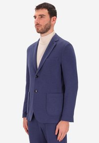 Blazer blu navy realizzato in un morbido tessuto, presenta due tasche frontali, due bottoni e un design con rever a punta. Indossato sopra un dolcevita color crema.