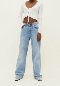 Person, der hellblaue weite Jeans, ein weißes, kurzärmeliges Oberteil mit langen Ärmeln und Knotenfront sowie metallische spitze Schuhe trägt, mit den Händen in den Taschen.