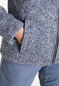 Gros plan d'une personne portant une veste en tricot bleu et blanc avec une main dans la poche avant et un pantalon bleu.