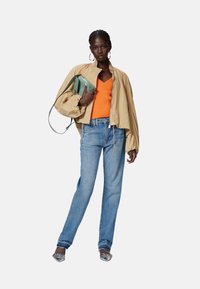 Giacca bomber beige con maniche voluminose, top lavorato a maglia arancione, jeans blu e scarpe con tacco argento. Tieni in mano una borsa verde.