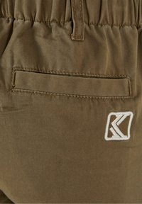 Gros plan sur une poche de pantalon en tissu marron avec une fente horizontale et un logo géométrique blanc brodé près du bas à droite.