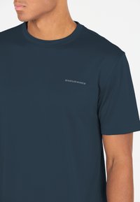 Man med marinblå kortärmad träningströja med texten "ENDURANCE" på vänster bröst, sedd från halsen till mitten av bålen.