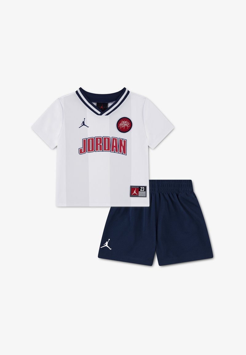 Tenue de basketball Jordan blanche et marine avec manches courtes, t-shirt col en V, logo rouge sur la poitrine, et short marine avec logo Jumpman.
