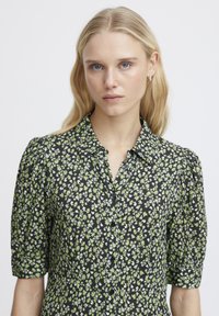 Blusa nera con motivo floreale bianco e verde, chiusura a bottoni sul davanti, colletto e maniche corte a sbuffo. Tessuto dalla consistenza liscia.