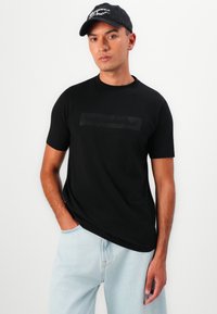 Svart T-skjorte med korte ermer laget av smooth stoff, med en matt logodesign på brystet. Sammen med lyseblå denim.