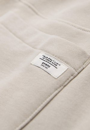 Beigenvärinen kangas, johon on ommeltu suorakulmainen valkoinen etiketti, jossa mustalla tekstillä lukee "YOUR ONLY LIMIT IS YOUR MIND" ja "OMBRE since 1989".