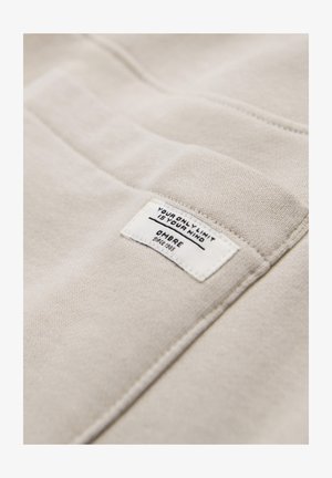 Beigenvärinen kangas, johon on ommeltu suorakulmainen valkoinen etiketti, jossa mustalla tekstillä lukee "YOUR ONLY LIMIT IS YOUR MIND" ja "OMBRE since 1989".