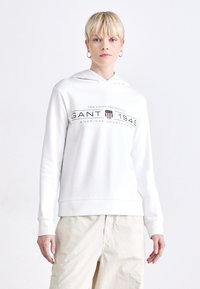 Hvit hettegenser med «GANT 1949» logo i mørk skrift, med ribbede mansjetter og en avslappet passform. Bæres med lyse beige bukser.