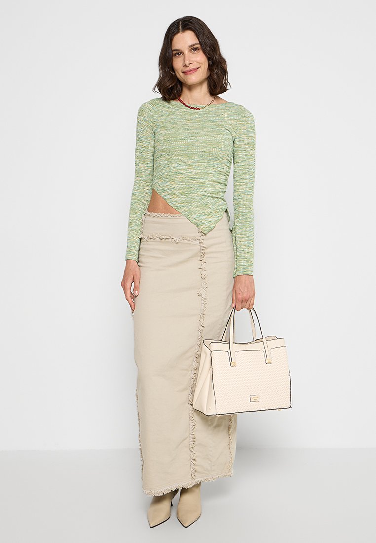 Groene gestructureerde langemouwen top met gedraaide zoom, gepaard met een beige maxi-rok met franjes. Ze houdt een crèmekleurige schoudertas vast.