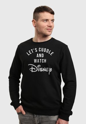Man met kort haar die een zwarte trui draagt met de tekst "Let's cuddle and watch Disney", kijkt opzij tegen een effen achtergrond.