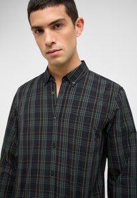 Junger Mann in einem schwarz-grünen karierten Hemd mit Brusttasche, vor einem schlichten hellgrauen Hintergrund stehend.