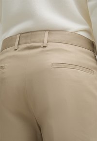 Pantaloni beige con una texture liscia, dotati di chiusura a pulsante e cerniera, e due tasche posteriori. La vita è fornita di passanti per la cintura.
