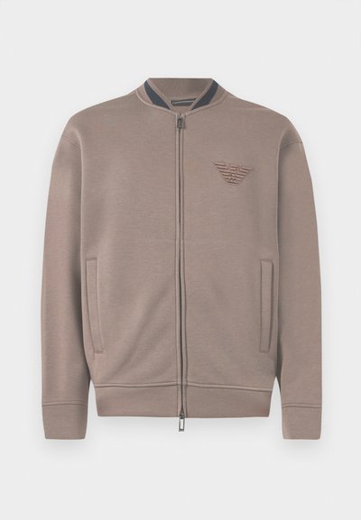 Šviesiai rudos spalvos zip-up džemperis su raštuotu apykakle, šoniniais kišeniais ir iškilmingu logotipu ant krūtinės. Minkšta medžiaga ir laisvas siluetas.