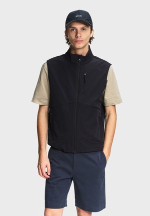 Jeune homme portant un gilet noir zippé par-dessus un t-shirt beige, un short bleu marine et une casquette noire avec le logo "AIGLE" sur un fond clair uni.