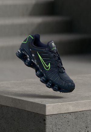 Tamsiai mėlynas „Nike“ bėgimo batelis su neoninės žalumos žyme ir akcentais, sklendžiantis virš betono paviršiaus, fone matyti pilkos laiptinės.