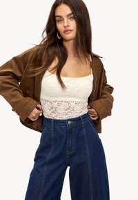 Chaqueta marrón con cremallera, blusa blanca de encaje y pantalones de mezclilla oscuros de tiro alto. El encaje texturizado y las costuras audaces acentúan el diseño del atuendo.