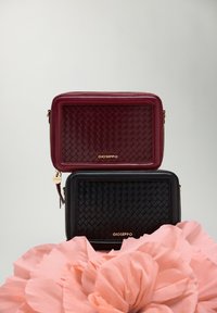 Deux pochettes en cuir tissé, une bordeaux et une noire, ornées de quincaillerie dorée et de pompons, reposant sur une grande fleur en papier rose.