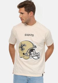Camiseta de algod�ón beige con un gráfico de un casco de fútbol americano de los New Orleans Saints en estilo vintage y la palabra "SAINTS" arriba en texto negro en negrita.