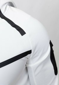 Haut de sport blanc avec une bande noire horizontale sur l'épaule et une poche zippée sur la manche. Texture lisse, design ajusté.