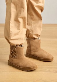 Bruine suède enkellaarzen met faux fur voering, voorzien van zijden vetersluiting en platte rubberen zolen, gecombineerd met beige broeken met elastische onderkant.