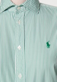Zeleno-bíle pruhovaná košile s límečkem, se zapínáním na knoflíky a malým zeleným logem na levé straně, vyrobená z lehkého materiálu.