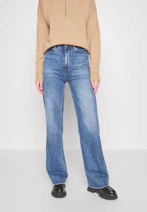 Personne portant un pull beige à fermeture éclair, un jean bleu taille haute à jambes larges et des chaussures noires épaisses, debout devant un fond uni.