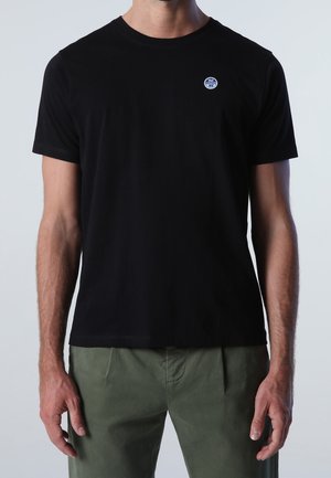 Sort bomuld t-shirt med korte ærmer, rund hals og en lille blå logo-patch på venstre bryst. Kombineret med grønne bukser.