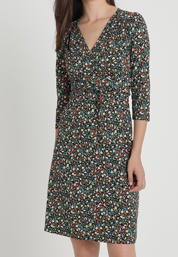 Femme portant une robe à fleurs longueur genoux, à manches longues, avec un décolleté en V et une taille cintrée, sur un fond sombre.