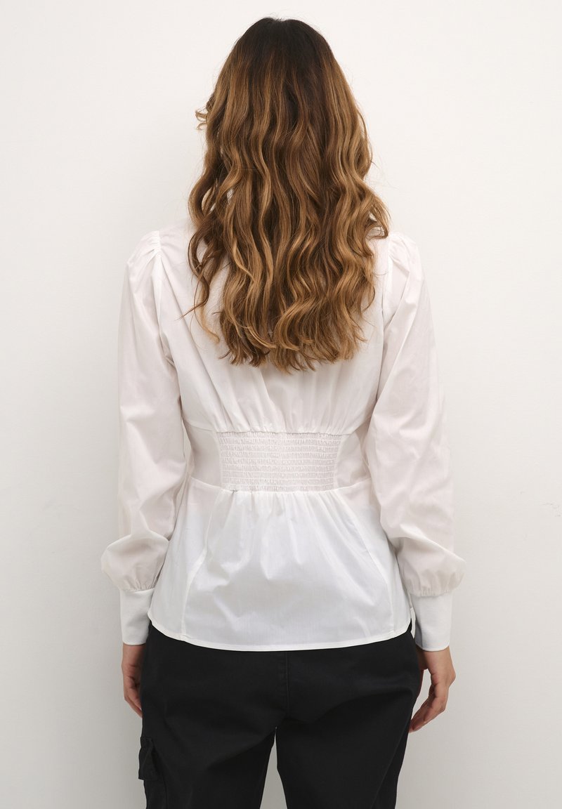 Eine Frau mit langen, gewellten braunen Haaren trägt eine weiße Bluse mit langen Ärmeln und schwarze Hosen und steht vor einer schlichten weißen Wand.