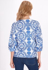 Blusa bianca con motivo floreale blu, maniche a tre quarti, scollatura rotonda e polsini arrotolati, caratterizzata da intricati disegni circolari.