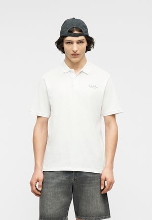 Νεαρός άνδρας με μαύρο καπέλο φορώντας ανάποδα, λευκό πόλο με κοντά μανίκια και γκρι τζιν σορτσάκι στέκεται μπροστά σε απλό φόντο.