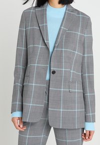 Szary marynarka w kratę z jasnoniebieskimi liniami, z zapięciem na jeden guzik, klapami w stylu notch oraz dwoma przednimi kieszeniami, noszona na jasnoniebieskim swetrze.