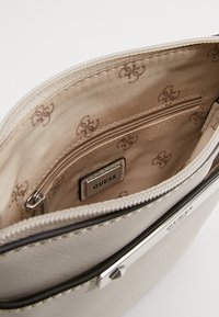 Bolso Guess beige que muestra el tejido interior con un patrón de logo marrón, un bolsillo con cremallera y una placa metálica de la marca Guess en el interior.