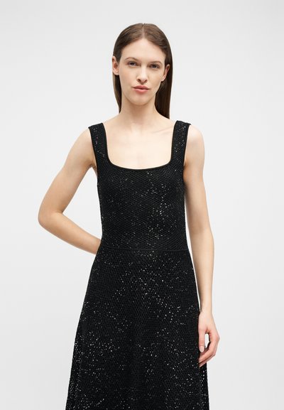 Γυναίκα με ίσια καστανά μαλλιά που φοράει αμάνικο μαύρο φόρεμα με παγιέτες και τετράγωνο ντεκολτέ, σε απλό ανοιχτό φόντο.