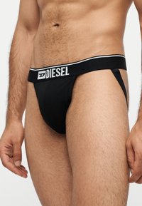 Mężczyzna noszący czarne męskie majtki typu jockstrap Diesel z widocznym pasem i górną częścią ud na neutralnym tle.
