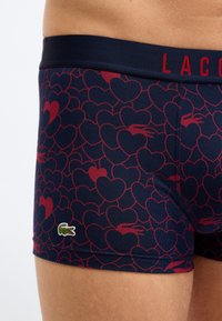 Σκούρες μπλε μποξεράκια με κόκκινα μοτίβα καρδιάς και μικρό πράσινο λογότυπο κροκοδείλου στο αριστερό μηρό, φορεμένα στο ισχίο ενός ατόμου.