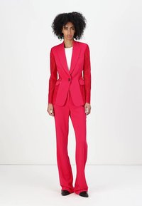 Crveno odijelo po mjeri s uskim sakoom i čipkastim rukavima, odgovarajuće hlače i elegantna silueta. Sjajne crne cipele zaokružuju izgled.