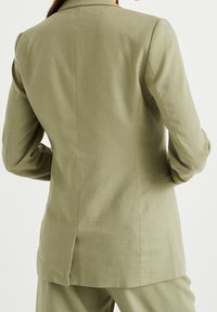 Olivgrön linneblazer med skräddarsydd passform, helkrage och slits bak. Har en slät yta och strukturerade axlar.