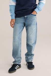 Világoskék farmernadrág, kopásos részletekkel, laza szabással és enyhén tapered (szűkített) lábbal. Fekete sportcipővel viselve, fehér csíkokkal.