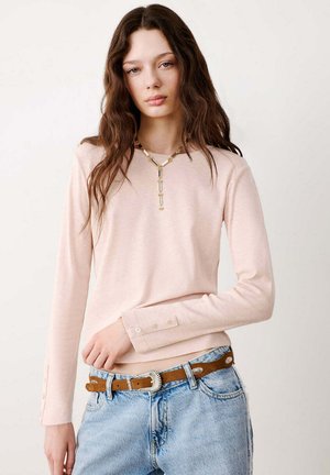 Jonge vrouw met lang bruin haar die een lichtroze top met lange mouwen, blauwe spijkerbroek, bruine riem en gouden hanger ketting draagt, staand tegen een witte achtergrond.