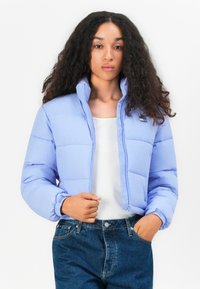 Lyseblå dunjakke med høy krage, glidelås foran og elastiske mansjetter. Brukt over en hvit skjorte og kombinert med mørke denimjeans.