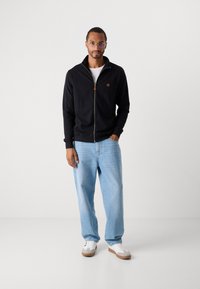 Hombre con gafas, chaqueta negra con cremallera, camiseta blanca, jeans de mezclilla azul claros y zapatillas blancas, de pie frente a un fondo blanco liso.