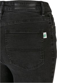 Pantalones vaqueros de mezclilla negra con una etiqueta de parche que dice "URBAN CLASSICS", una etiqueta de hoja verde en la parte trasera y diseño de bolsillos estándar. Textura suave.