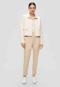 Veste blanche raccourcie avec deux poches avant, associée à un pantalon beige clair ajusté et des baskets blanches. Tissu lisse, design minimaliste.