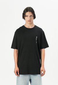 Schwarzes Baumwoll-T-Shirt mit kurzen Ärmeln, versehen mit weißem Text auf der linken Seite. Einfacher, lässiger Schnitt; kombiniert mit hellblauen Jeans.